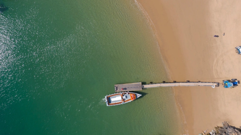 Drone shot of a golden sanded beach from above. There's a red boat moored by a jetty.